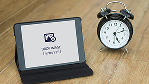 Black iPad image mockup of an Apple tablet on a stand beside an alarm clock on a wooden surface, 1470 x 1151